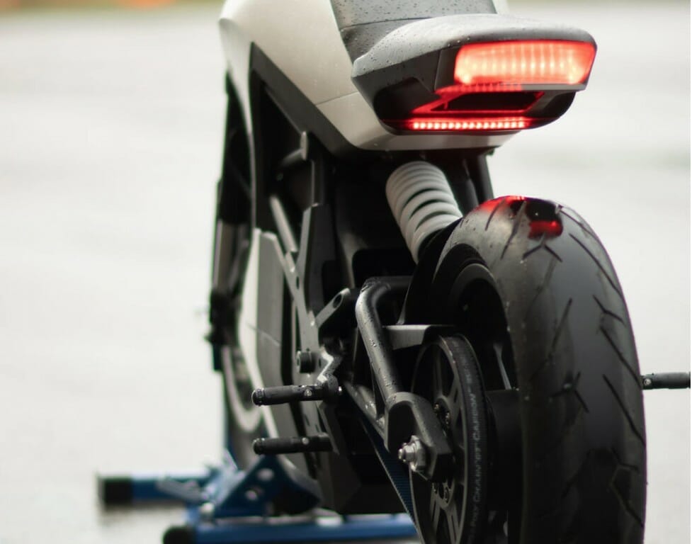 Close-up view of an electric motorcycle's rear wheel, suspension, and LED tail light, positioned on a stand in an outdoor setting.