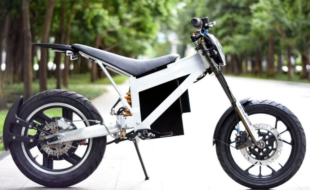 Electric motorcycle parked on a paved path surrounded by green trees. The motorcycle has a sleek design with large tires and modern components.