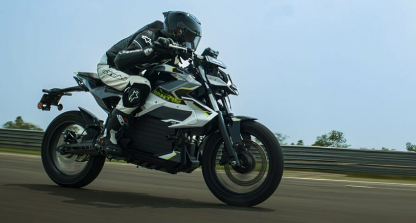 A motorcyclist in full gear rides a black and white electric motorcycle on a paved road with a guardrail in the background.