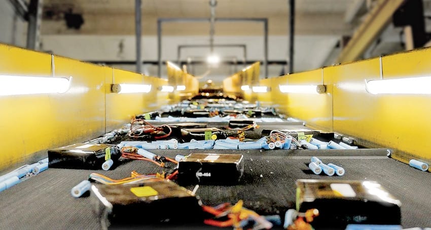 Conveyor belt in a factory carrying electronic components and connected wires, with yellow side rails and overhead lighting.