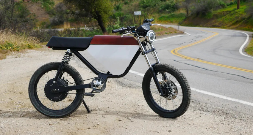 An electric bike is parked on a gravel area beside a winding road with a green hillside in the background.