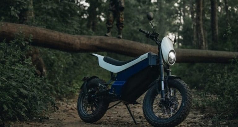 A blue and black electric motorcycle is parked on a dirt path in a forest, with a large fallen tree in the background and a person standing on the tree trunk.