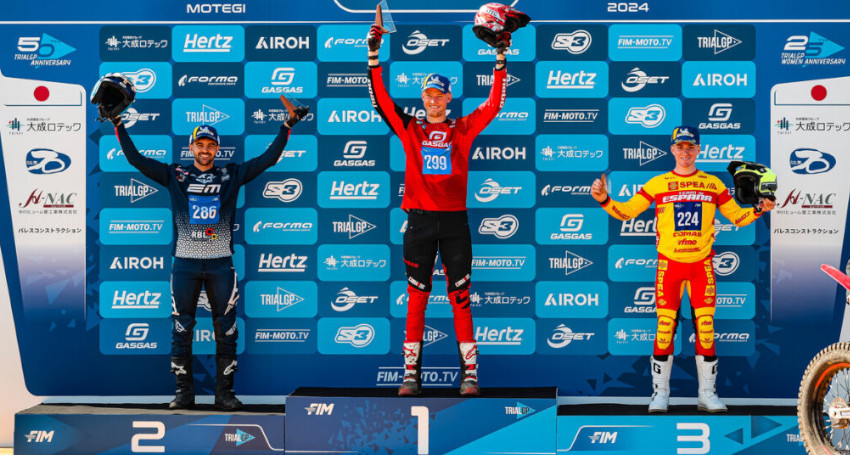 Three motocross riders stand on a podium, celebrating their victories at the 2024 FIM TrialGP World Championship. The center rider in red is in first place, the left rider in blue is in second place, and the right rider in yellow is in third place.