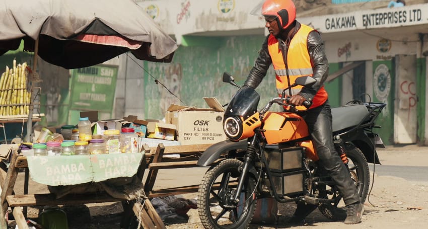 A person in an orange helmet and vest sits on an orange motorcycle by a street stall with items and jars on display.