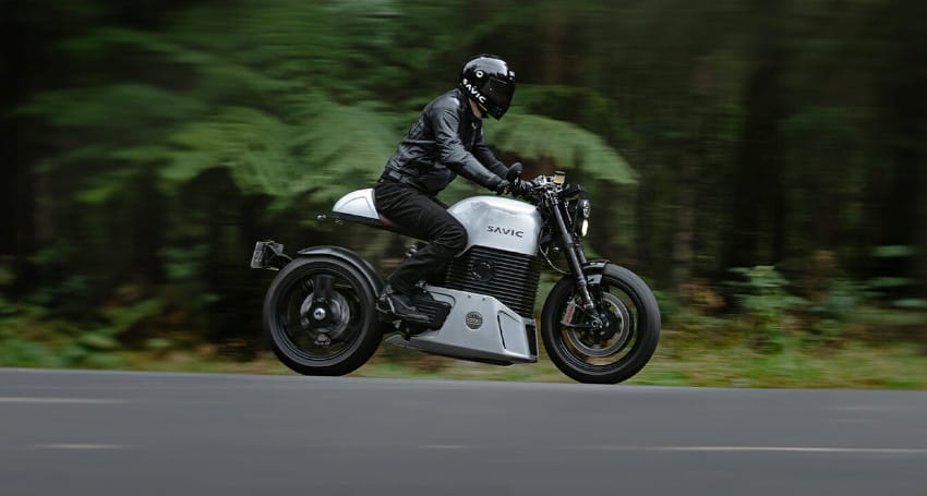 A person wearing black leather gear and a helmet rides a modern electric motorcycle on a paved road with blurred greenery in the background.