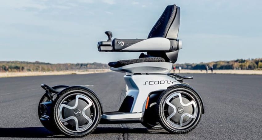 A modern, four-wheeled mobility scooter is shown on an empty road with a clear sky and trees in the background. The scooter has a sleek design and appears to be designed for enhanced accessibility.