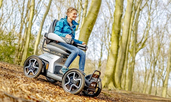 A woman riding a Scoozy in a wooded area.