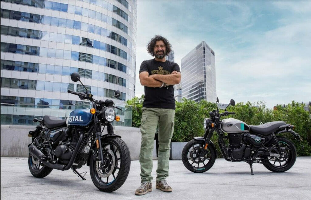 A person with crossed arms stands outdoors between two modern motorcycles, with tall buildings in the background.