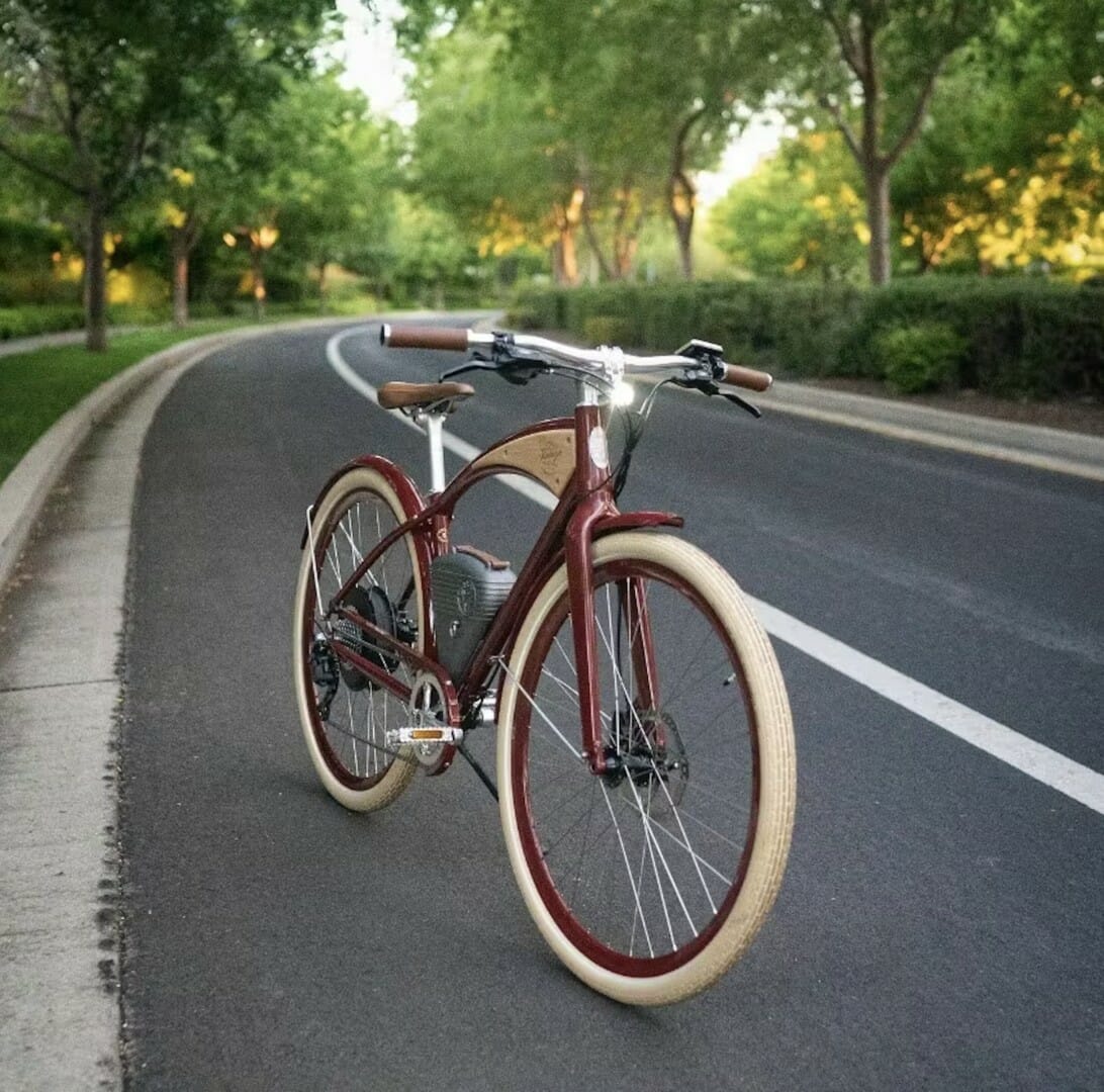 A vintage-style bicycle with beige tires is parked on a paved road surrounded by trees and greenery.