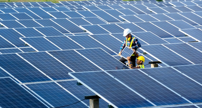 Two workers in safety gear are installing solar panels in a large solar farm, with rows of panels visible under a clear sky.