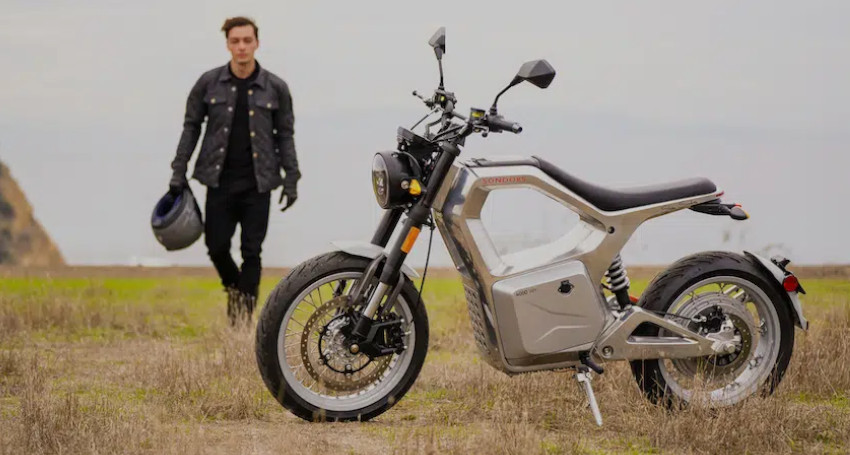 A person in a black jacket walks holding a helmet towards a parked grey electric motorcycle in an open, grassy area.