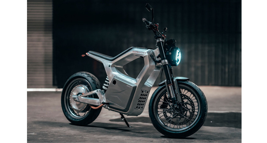 A silver electric motorcycle with a minimalist design featuring a circular headlight and black tires, parked indoors on a concrete floor.