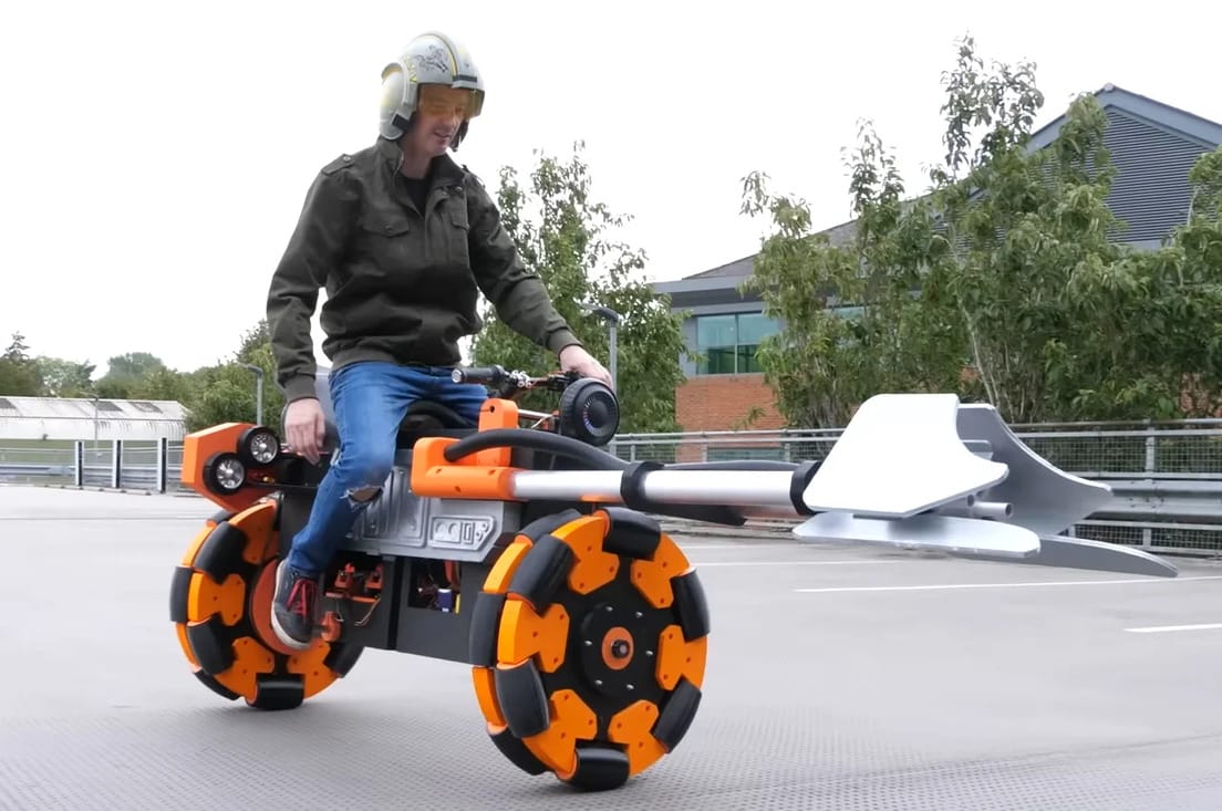 Person wearing a helmet and jacket riding a uniquely designed motorcycle-like vehicle with large orange and black wheels and an extended shovel at the front, in an outdoor setting.