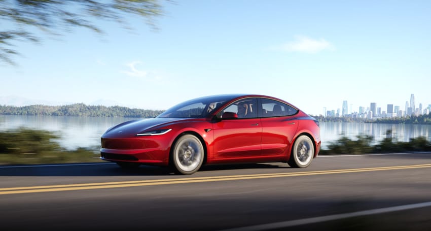 Red electric car driving on a lakeside road with a city skyline in the background.