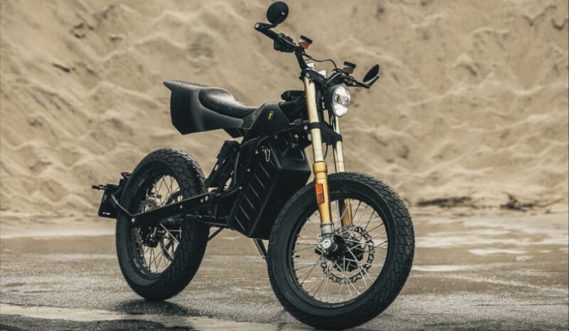 A black electric motorcycle is parked on a wet, sandy surface with a large sand pile in the background.