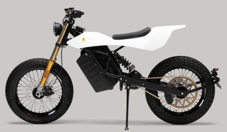 Side profile of a white electric motorcycle with a minimalist design, featuring a large black battery pack, golden front suspension, and off-road tires.