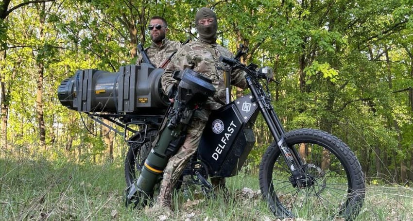 Two individuals in military gear stand outdoors; one is on a Delfast e-bike equipped with a large weapon, amidst a wooded area.