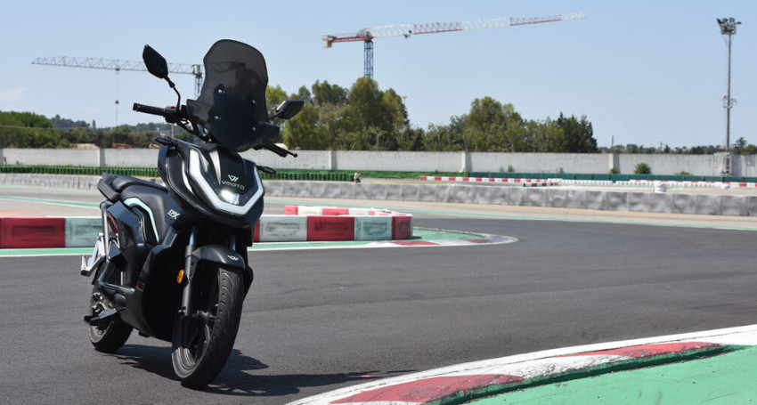 A black and silver scooter with a superfast charging battery is parked on an empty racetrack with red and white barriers, and construction cranes are visible in the background.