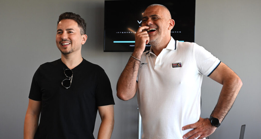 Jorge Lorenzo and Graziano Milone stand together, one in a black shirt and the other in a white polo shirt, both smiling and looking in the same direction. A monitor displaying a logo for a Superfast Charging Battery is visible in the background.