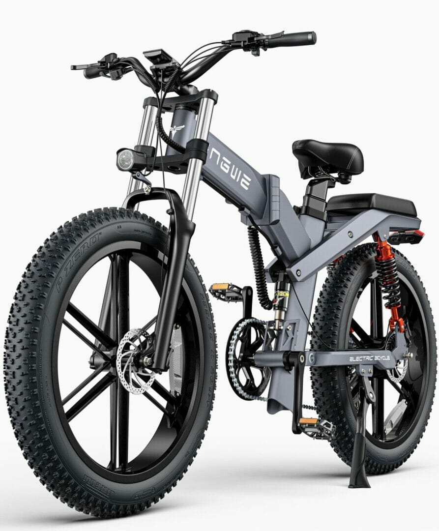 A gray electric mountain bike with large knobby tires, a front suspension fork, dual shock absorbers at the rear, and a mounted battery on the frame. The bike is on a stand.