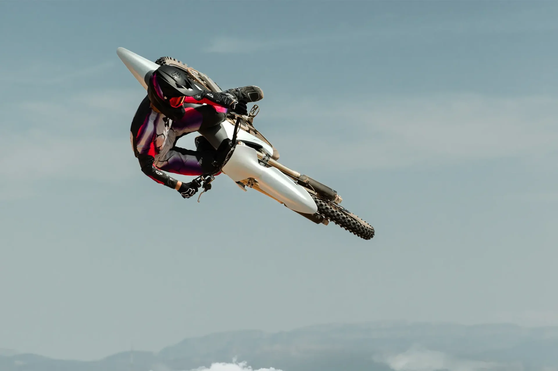 A motocross rider in mid-air performing a jump against a clear sky background. The rider is dressed in a black, white, and pink outfit. The motorcycle is tilted to the side.