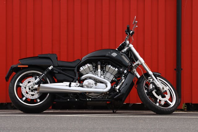 Black motorcycle parked in front of a red corrugated metal wall on a paved surface.