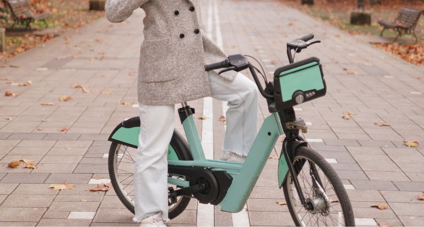 Person in a gray coat and white pants stands with one foot on a green rental bike on a paved, leaf-strewn path in a park.