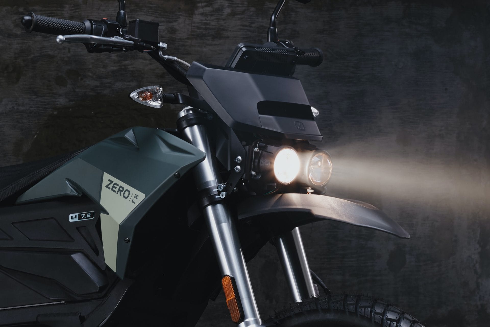 Close-up of a dark-colored motorcycle with headlights on against a dark background. The motorcycle features the brand name "Zero" and model details "FX" and "7.2".