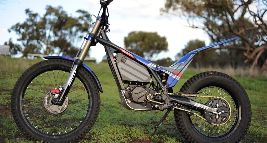 A blue and black electric dirt bike is parked on a grassy field with trees in the background.