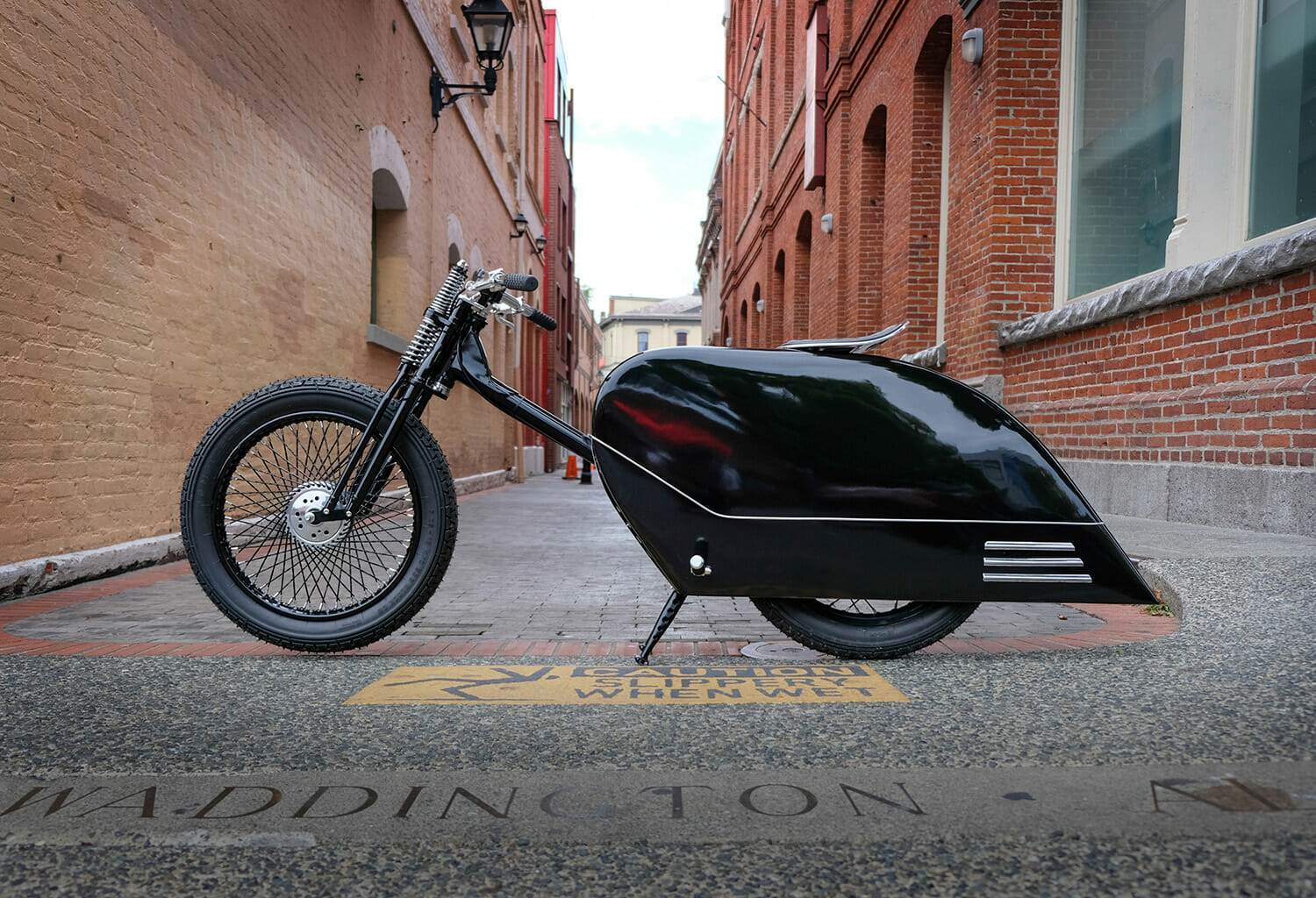 A sleek black electric motorcycle is parked in a narrow alleyway between brick buildings. The bike features a unique aerodynamic design with a streamlined body and large front wheel.