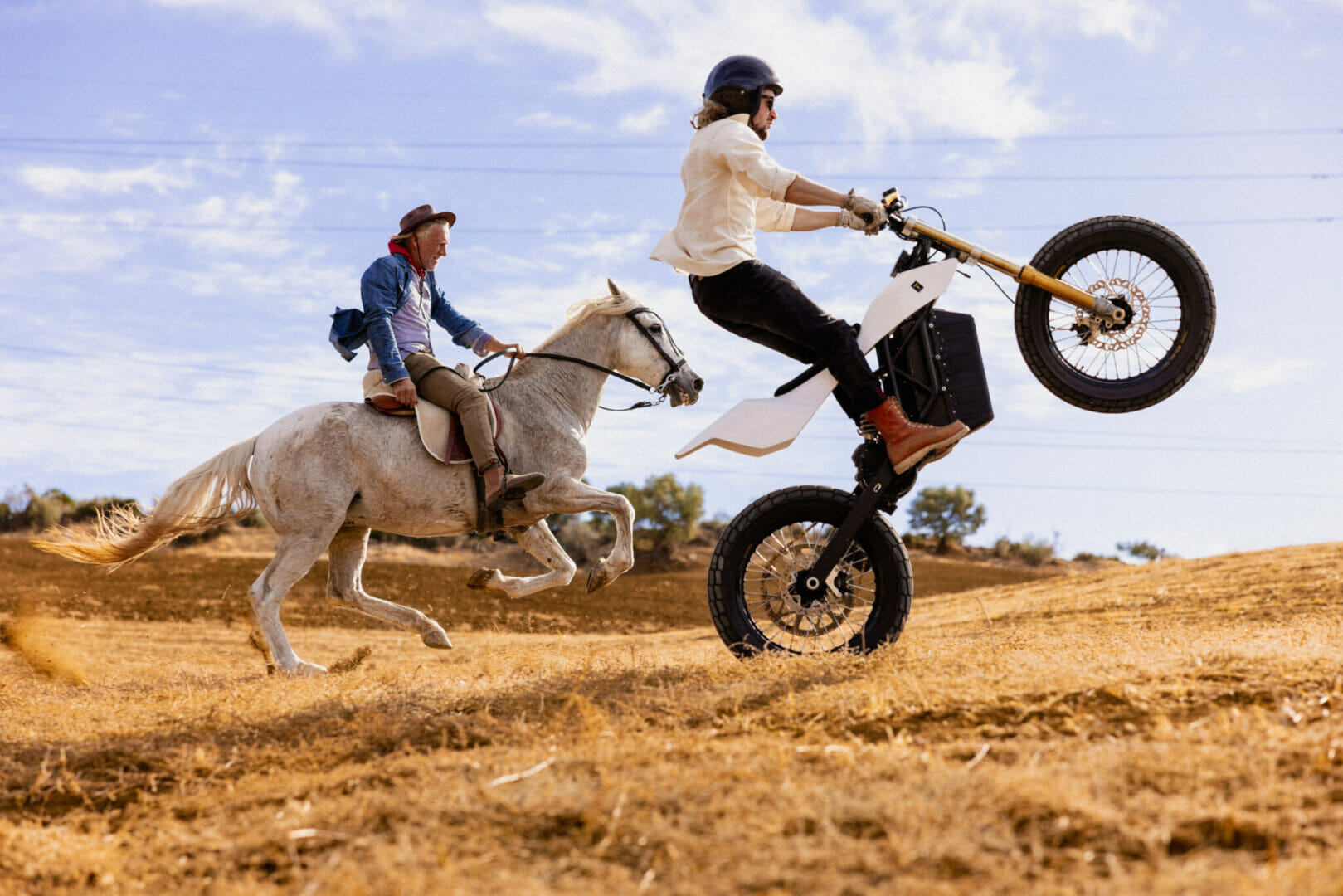 Person riding a wheelie on a dirt bike while another person rides a galloping white horse in a dry, hilly landscape.