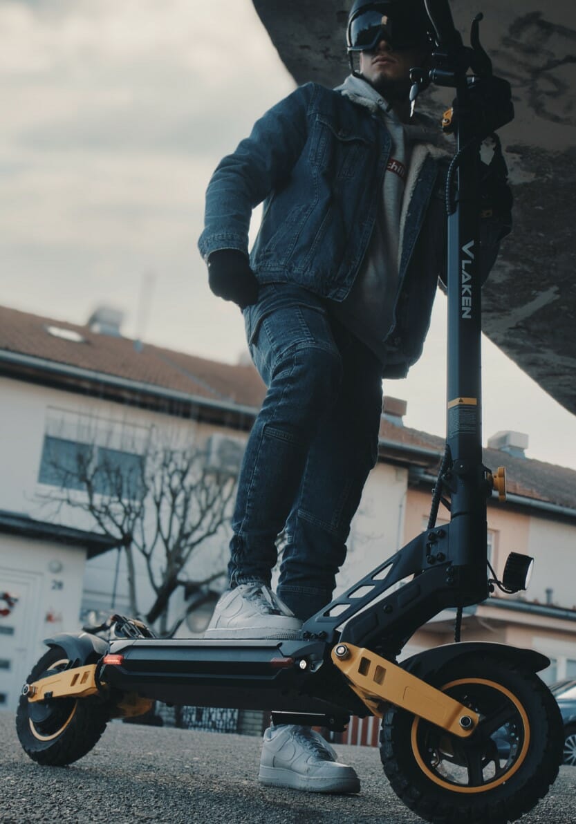 Person wearing a blue denim jacket and helmet stands on a black and yellow electric scooter under a bridge. Residential buildings and a leafless tree are visible in the background.