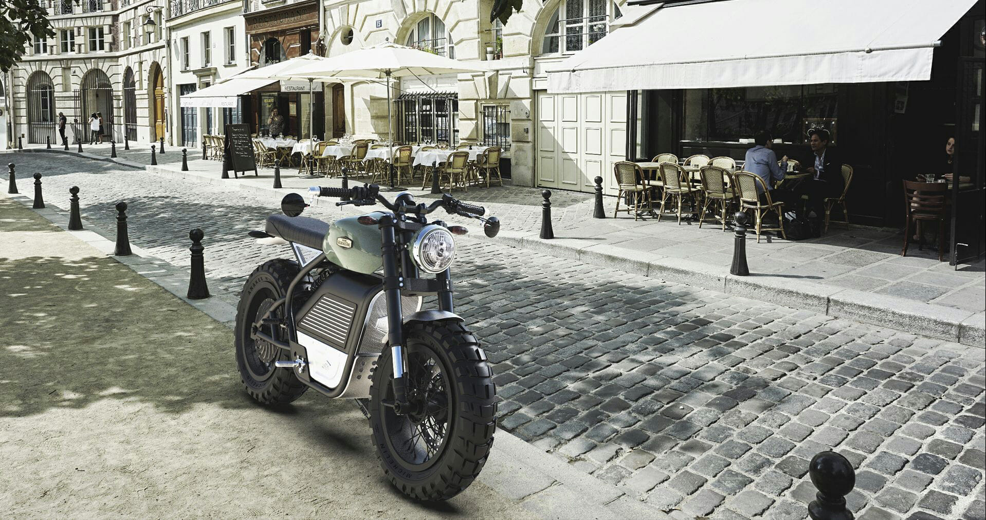 A motorcycle is parked on a cobblestone street next to a café with outdoor seating. People are dining at the café, and surrounding buildings have a classic architectural style.