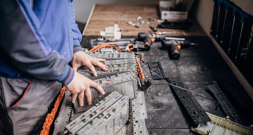 Person in a blue jacket assembles or repairs an electrical device on a workbench, surrounded by tools and cables.