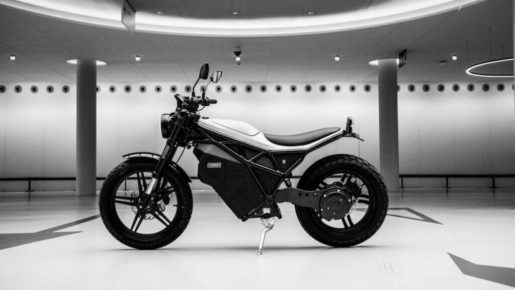 A black and white image of an electric motorcycle displayed in a modern, well-lit showroom with circular lights on the ceiling and columns in the background.