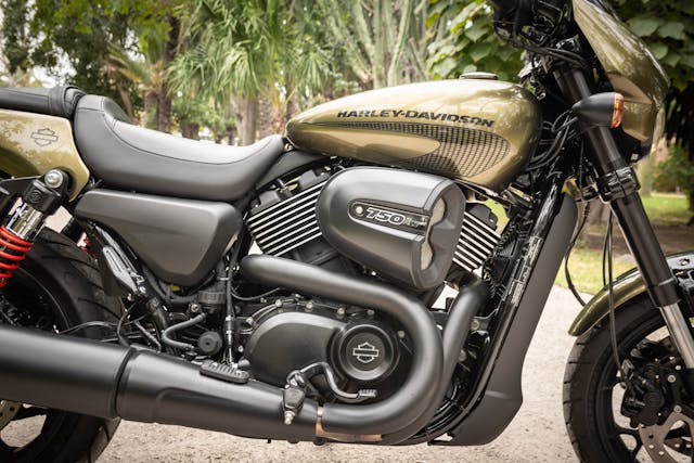 Close-up view of a Harley-Davidson motorcycle, highlighting the engine, exhaust, and branded fuel tank, with greenery visible in the background.
