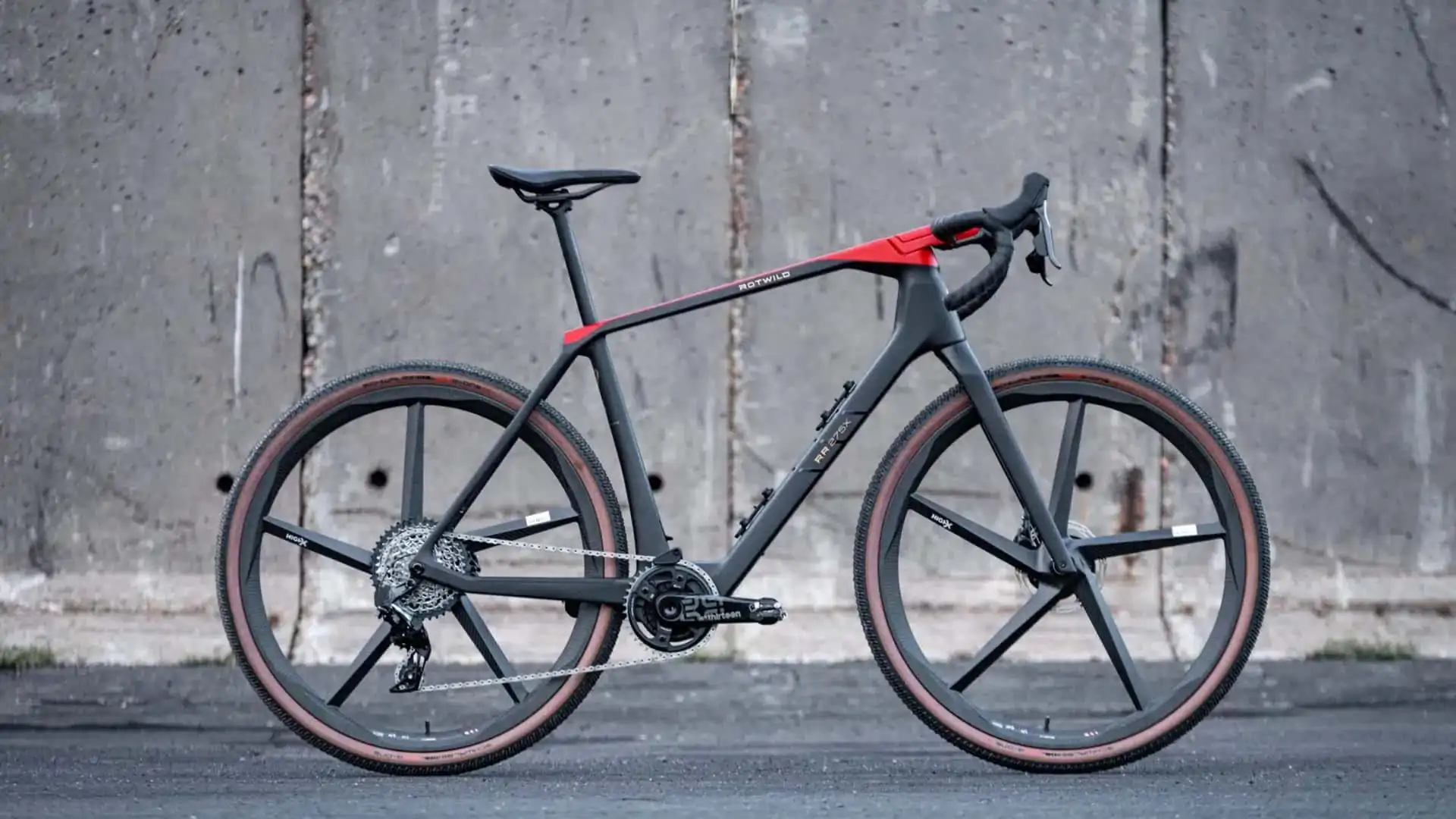 A sleek black and red road bike stands upright against a concrete wall, featuring thick tires and aerodynamic frame design.