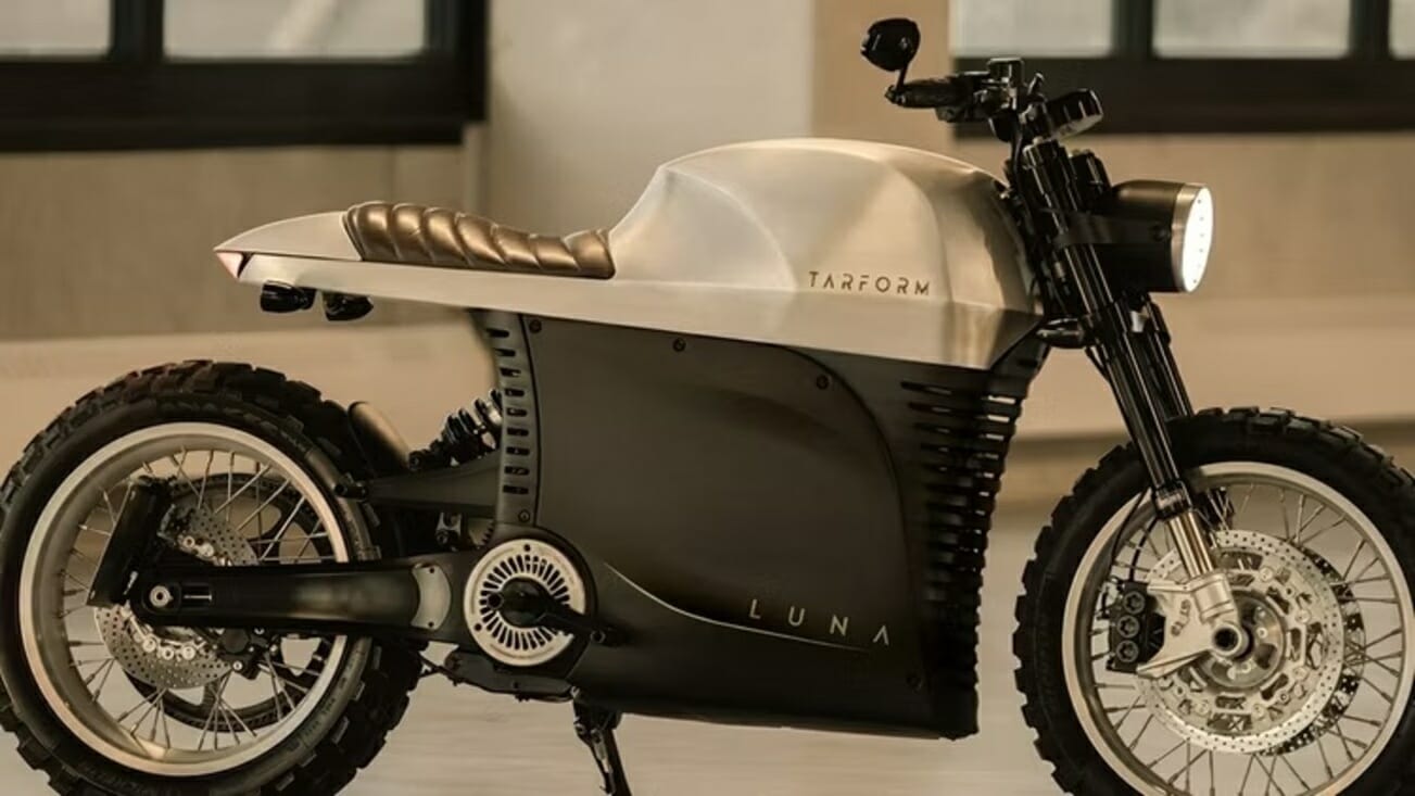 A silver and black motorcycle labeled "Tarform Luna" with a sleek design, large tires, and a minimalist seat, positioned indoors with windows in the background.