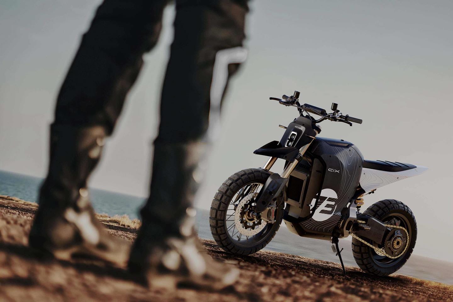 A person in black motorcycle gear stands near a futuristic electric motorcycle on a dirt road near the sea.