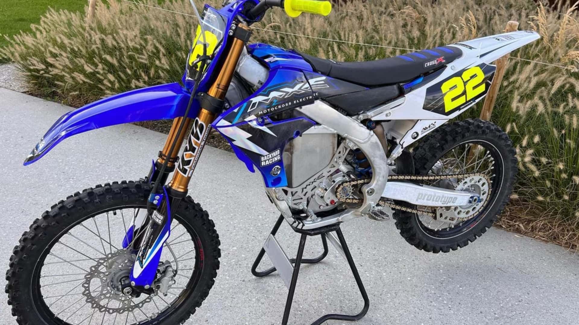 A blue and white dirt bike with the number 22 displayed on its design sits on a stand in front of tall grass.