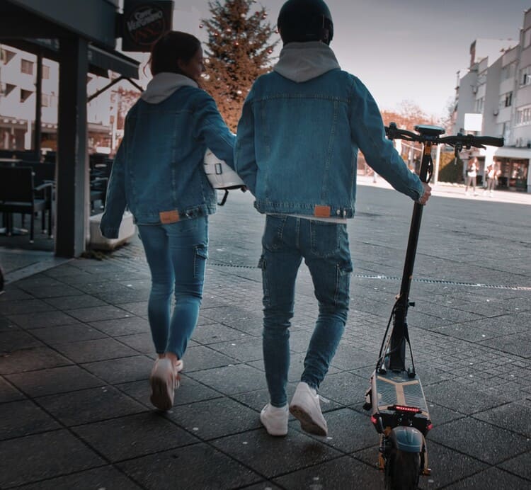 Two individuals in denim outfits walk holding hands; the person on the right is also walking a scooter. The scene is set in an urban area with buildings in the background.