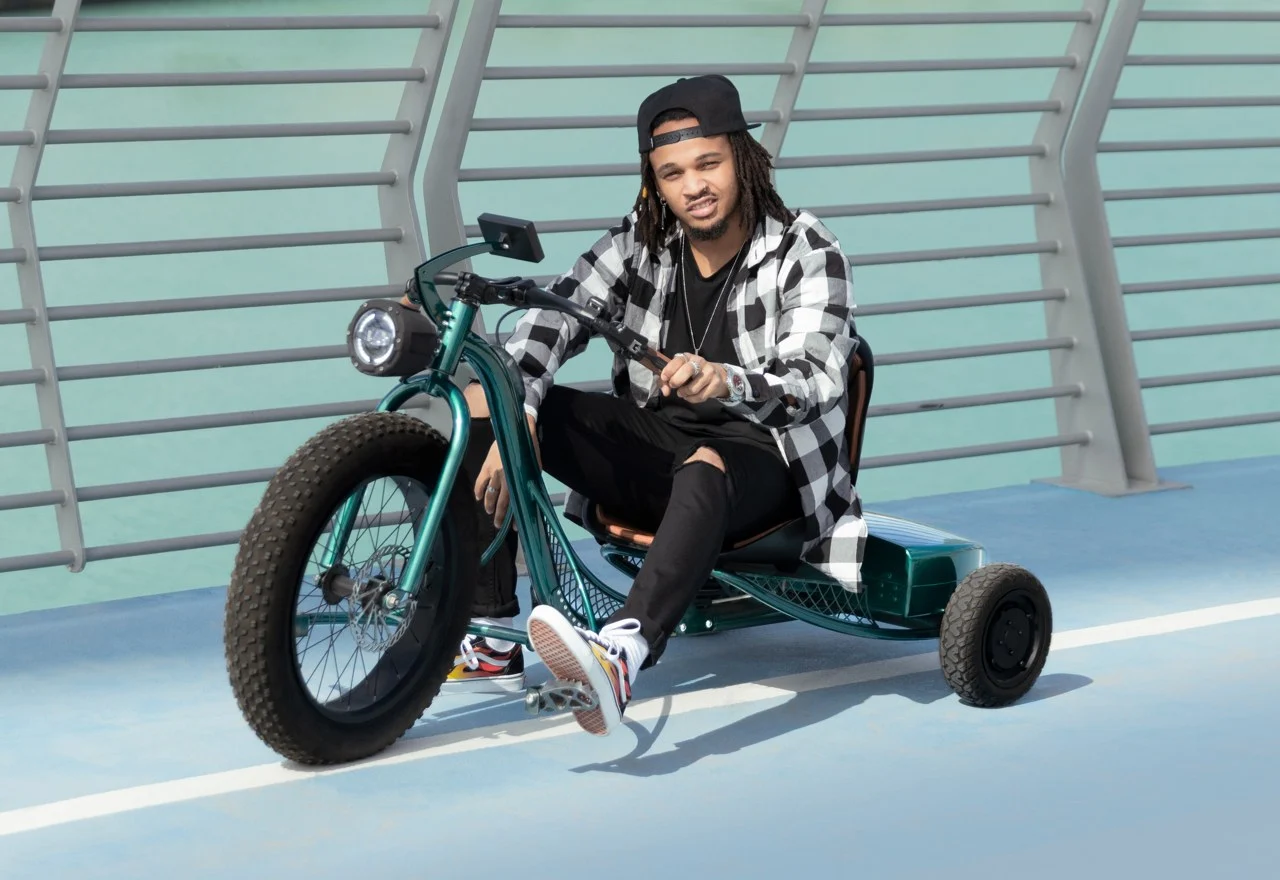 A person wearing a cap, black and white flannel shirt, and black jeans sits on a green tricycle with large front wheels and two smaller rear wheels. The background features a green metal wall.